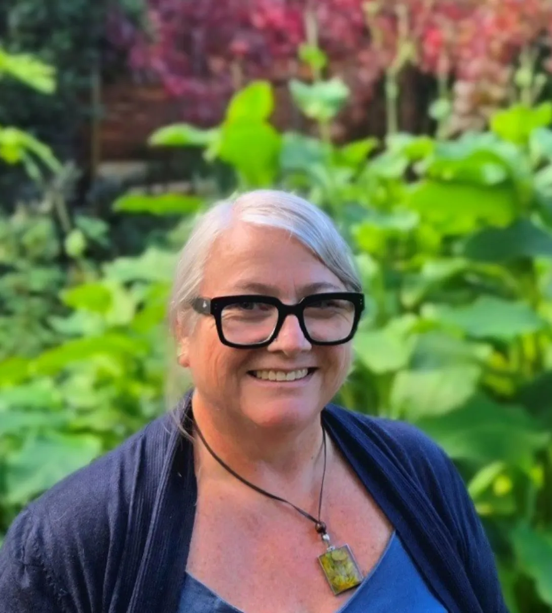 Smiling woman with grey hair and bold black glasses wearing a navy cardigan in a garden.