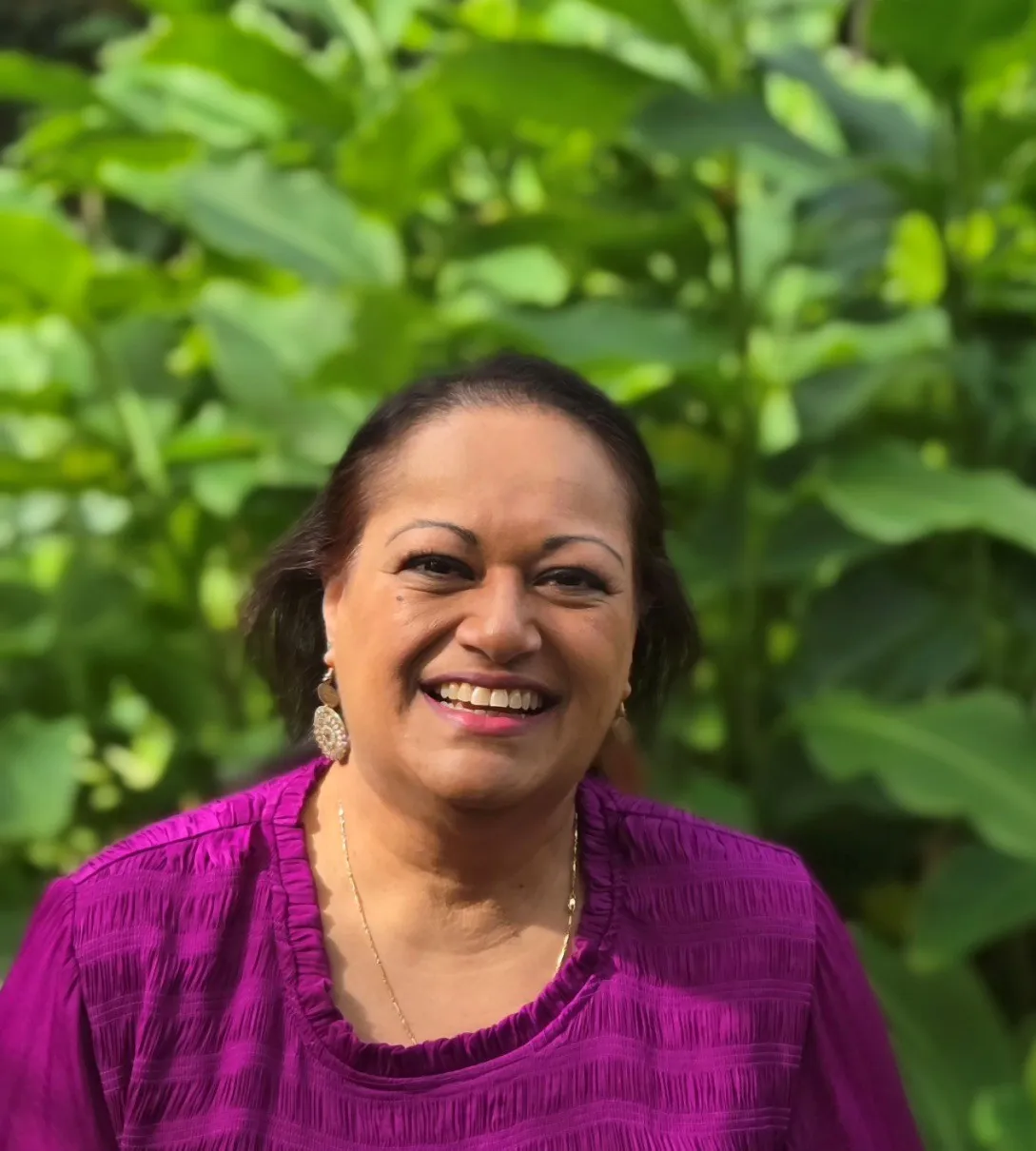 Smiling woman in a textured purple shirt standing in front of lush green foliage.