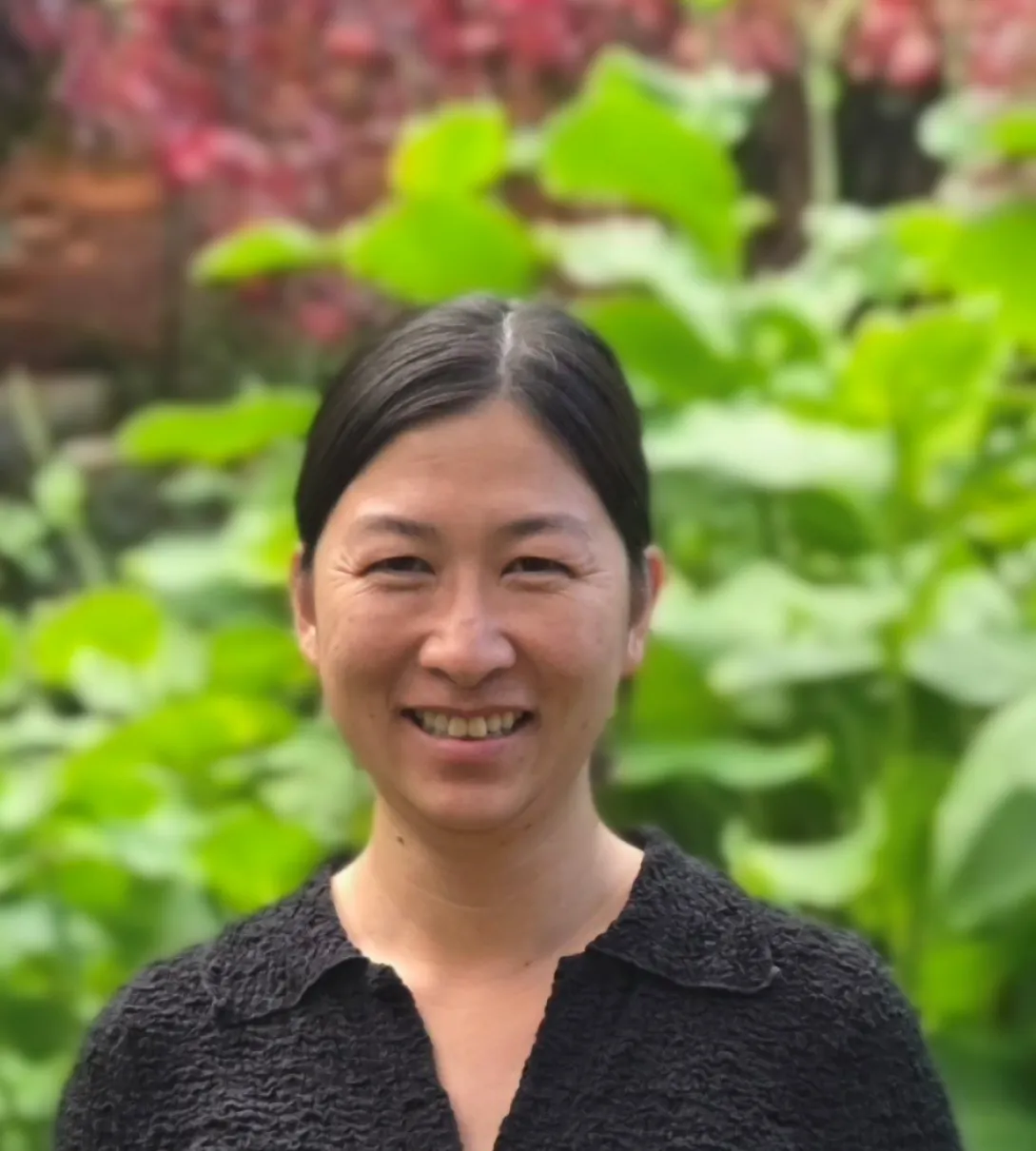 Smiling woman with dark hair wearing a black textured shirt in front of a green garden background.