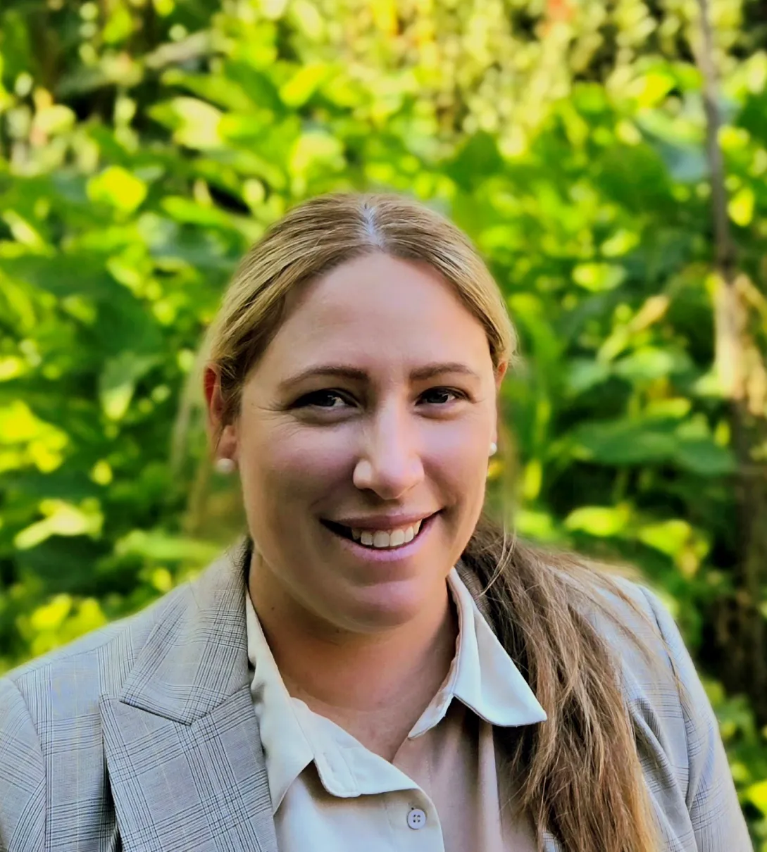 Smiling woman with blonde hair wearing a grey plaid blazer outdoors against a green leaf background.
